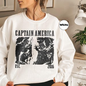 May include: White sweatshirt featuring a black graphic of Captain America with the text "CAPTAIN AMERICA" above the image and "EST. 2016" below. The sweatshirt is a crew neck style.
