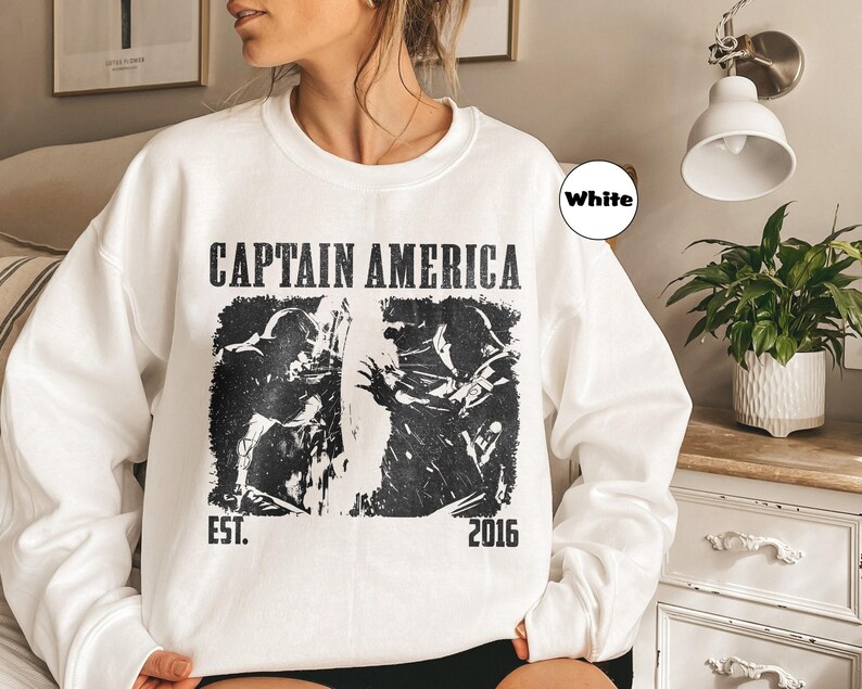 May include: White sweatshirt featuring a black graphic of Captain America with the text "CAPTAIN AMERICA" above the image and "EST. 2016" below. The sweatshirt is a crew neck style.