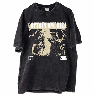 May include: Black t-shirt with a distressed graphic featuring the text "CAPTAIN AMERICA" above a beige image of two figures in action. The text "EST. 2016" is below the image. The shirt has a vintage, worn look.