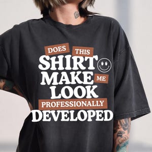 Könnte beinhalten: Ein anthrazitfarbenes Kurzarm-T-Shirt mit weißem Text DOES THIS SHIRT MAKE ME LOOK PROFESSIONALLY DEVELOPED, braunen rechteckigen Akzenten und einem weißen Umriss eines lächelnden Gesichts auf der rechten Seite des Textes.