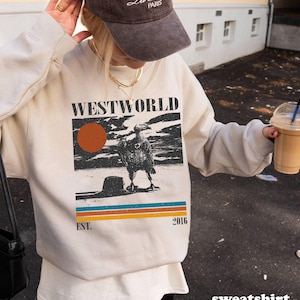 May include: Cream-colored sweatshirt with a graphic featuring the word "WESTWORLD", a sun, and a bird. Below the graphic are horizontal stripes in blue, orange, and yellow. The text "EST. 2016" is also visible. A brown cap is worn.