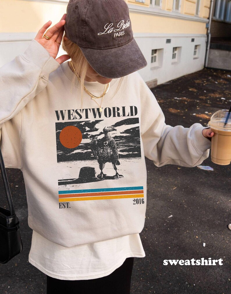 May include: Cream-colored sweatshirt with a graphic featuring the word "WESTWORLD", a sun, and a bird. Below the graphic are horizontal stripes in blue, orange, and yellow. The text "EST. 2016" is also visible. A brown cap is worn.