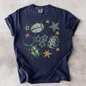 Op de afbeelding: Een donkerblauw t-shirt met korte mouwen en een grafische print van twee groene kevers en verschillende gouden en witte sterren. Het ontwerp bevat een grote groene kever met opvallende kaken en een kleinere gevlekte kever.