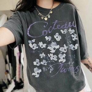 May include: A charcoal gray short-sleeve t-shirt featuring a purple script logo that reads Cocteau Twins. The front of the shirt is decorated with a repeating pattern of white, stylized floral illustrations scattered across the chest area.