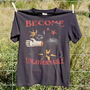 Może przedstawiać: Ciemnoszary t-shirt z krótkim rękawem z grafiką przedstawiającą czarnego kota trzymającego w pysku odciętą dłoń. Nad kotem znajduje się napis "BECOME", a pod nim "UNGOVERNABLE", w otoczeniu ilustracji żółtych i czerwonych gwiazd.