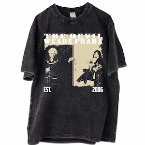 Könnte beinhalten: Schwarzes T-Shirt mit einem verblichenen Grafikdesign aus dem Film "Der Teufel trägt Prada". Das Design zeigt zwei Frauen und den Text "THE DEVIL WEARS PRADA" darüber, mit "EST. 2006" darunter. Das Shirt hat einen Vintage-Look.