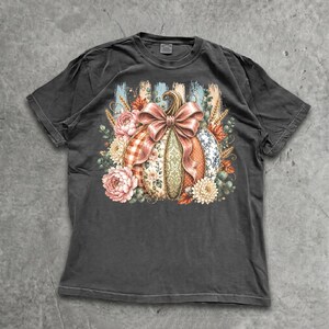 Może przedstawiać: Ciemnoszary t-shirt z krótkim rękawem z centralną grafiką przedstawiającą dynię ozdobioną dużą różową kokardą. Dynia jest otoczona elementami kwiatowymi, kłosami pszenicy i liśćmi w odcieniach różu, pomarańczu, zieleni i kremu.