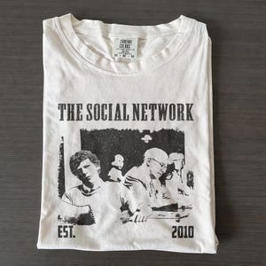 May include: White t-shirt with a black and white graphic from the movie "The Social Network." The shirt features the film's title, the year 2010, and a black and white image of the movie's characters. The shirt is folded and has a tag.