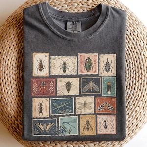 Può includere: Una t-shirt grigio antracite a girocollo con una stampa grafica di quindici francobolli in stile vintage, ognuno dei quali mostra una diversa illustrazione di insetti, tra cui coleotteri, farfalle, falene e libellule con vari valori.
