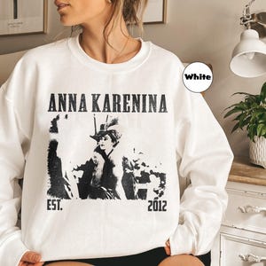 Puede incluir: Sudadera blanca con el texto "ANNA KARENINA" en negro, junto con un gr&aacute;fico en blanco y negro de una mujer con sombrero. La sudadera tambi&eacute;n presenta el texto "EST. 2012".