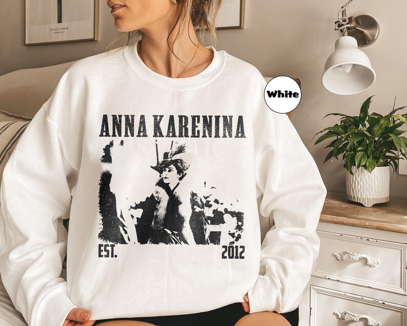 Puede incluir: Sudadera blanca con el texto "ANNA KARENINA" en negro, junto con un gr&aacute;fico en blanco y negro de una mujer con sombrero. La sudadera tambi&eacute;n presenta el texto "EST. 2012".