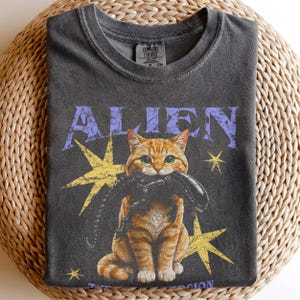 Puede incluir: Una camiseta gris carbón con el gráfico de un gato atigrado naranja que sostiene una criatura alienígena negra en la boca. La palabra ALIEN está impresa en letras moradas desgastadas sobre el gato, rodeada de estrellas amarillas.