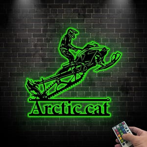 May include: Black metal wall art depicting a snowmobile with a rider, illuminated by green neon light. The text "Arctic cat" is displayed below the snowmobile. The art is mounted on a dark brick wall.