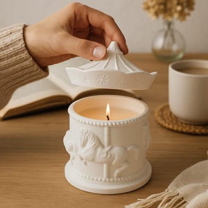 Op de afbeelding: Een witte kaars met carrouselthema en een brandende lont. De kaarsenhouder van keramiek heeft reliëfdetails van carrouselpaarden. Het deksel, vastgehouden door een hand, heeft de vorm van een carrouseldak met steruitsparingen.