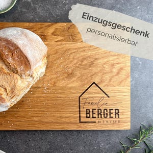 May include: A wooden chopping board with a loaf of bread on top. The board features a house graphic with the name "Berger" and coordinates. The text "Einzugsgeschenk personalisierbar" is in the upper right. The bread is light brown with a white dusting.