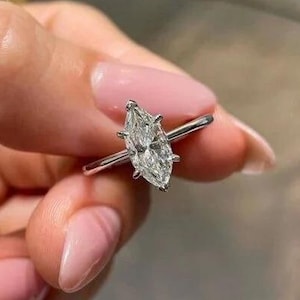 May include: A marquise-cut diamond ring with a thin silver band. The diamond is set with prongs and is held up by a silver band. The ring is held up by a person's hand with pink nail polish.