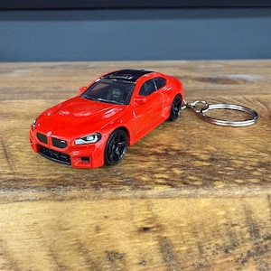 May include: A red toy car keychain with a black roof and black wheels. The miniature car has a detailed front grille and headlights, attached to a silver keyring. The car is sitting on a wooden surface.