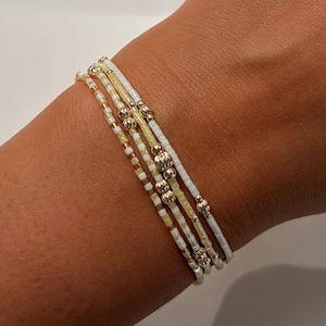May include: A set of four stacked bracelets featuring small white and pale yellow seed beads. The bracelets are accented with small, round, metallic gold-toned beads of varying sizes, including smooth spheres and textured, faceted gold-toned beads.