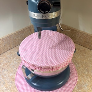 Op de afbeelding: Een blauwe KitchenAid-keukenmixer met een roze en witte geruite komdeksel en bijpassende onderzetter. De mixer heeft een zilveren afwerking en het KitchenAid-logo. De komdeksel heeft een gerimpelde rand.