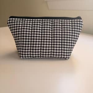May include: A quilted pouch featuring a black and white gingham check pattern. The rectangular bag has vertical stitching lines across the fabric and is secured at the top with a black zip closure that runs along the entire length of the opening.