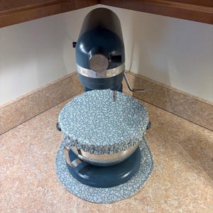 May include: A teal KitchenAid stand mixer with a floral patterned bowl cover and base cover. The mixer is positioned on a countertop in a kitchen setting. The bowl cover and base cover have a light blue background with white floral designs.