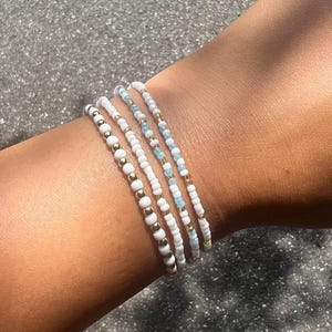 May include: Four beaded bracelets in white, gold, and light blue are stacked on a wrist. The bracelets feature small, round beads in varying patterns. The gold beads add a touch of shine, while the white and blue beads create a delicate, summery look.