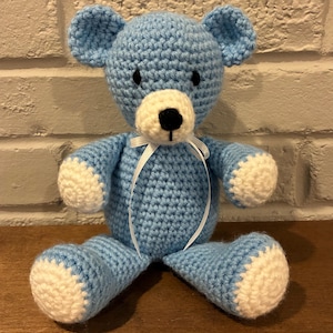 May include: A light blue crocheted teddy bear with a white muzzle, white paw pads, and black embroidered eyes and nose. The bear features a thin white ribbon tied in a bow around its neck, with a textured yarn construction throughout its body.