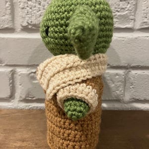 以下が含まれることがあります： 人気のSFフランチャイズのキャラクターに似せた手作りのかぎ針編み人形。人形は、大きな耳のある緑色の頭、ベージュのスカーフ、茶色の体を持っています。人形は、きついかぎ針編みのステッチで作られています。