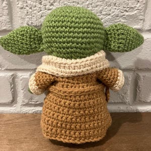 以下が含まれることがあります： 大きな耳を持つキャラクターの手作りの編み人形。茶色のローブとクリーム色の襟を着用しています。人形の頭と耳は緑色で、体は茶色です。人形の高さは約15cmです。