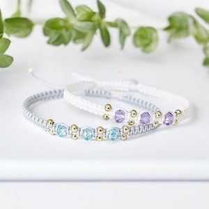 May include: Two friendship bracelets with white cord and small beads. One bracelet has light blue beads and the other has purple beads.