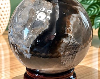 May include: A polished spherical stone with a mix of dark brown, black, and translucent gray patterns, featuring a small natural cavity at the top and a white circular graphic design on the surface, resting on a dark brown wooden circular stand.