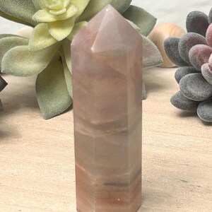 May include: A tall, hexagonal-shaped crystal tower in shades of pink and tan. The polished stone has a pointed top and visible natural banding. The crystal is displayed with artificial succulents in the background.