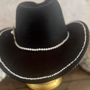 May include: A black cowboy hat with a pearl beaded trim around the brim and crown. The hat has a classic western style with a slightly curved brim and a rounded crown. A small feather is attached to the side of the hat.