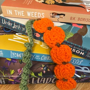 May include: A handmade crochet bookmark featuring four orange pumpkins with brown stems and a green vine. The bookmark is placed on a stack of books, including one titled "In The Weeds" by B.K. Borison.