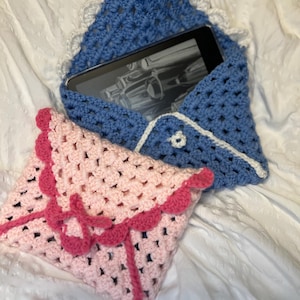 May include: Two handmade crochet cases, one blue and one pink, rest on a white surface. The blue case, with a white lace trim, holds an e-reader. The pink case features a scalloped edge and a pink tie closure. Both cases are made with a granny stitch.