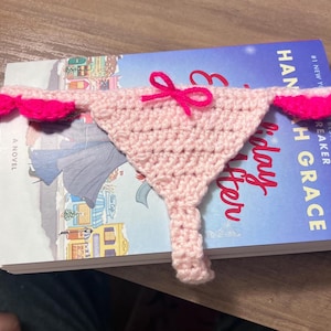 Peut inclure: Marque-page en crochet rose clair fait à la main, en forme de sous-vêtements, avec un petit nœud rose. Les bords sont ornés de détails festonnés rose vif. Le marque-page est posé sur un livre.