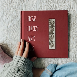 May include: A square, deep red hardback book with the white text "HOW LUCKY ARE WE" printed on the front cover. A vertical strip of four black and white photo booth pictures showing two people embracing is attached to the right side of the cover.