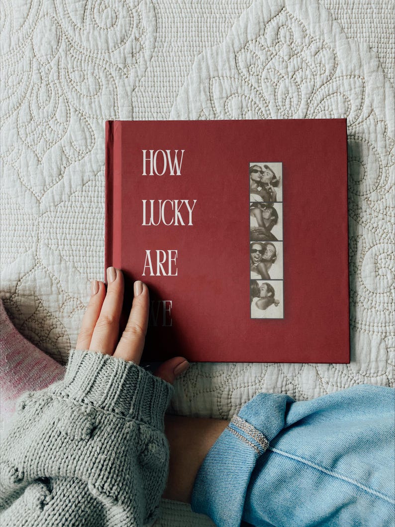 May include: A square, deep red hardback book with the white text "HOW LUCKY ARE WE" printed on the front cover. A vertical strip of four black and white photo booth pictures showing two people embracing is attached to the right side of the cover.