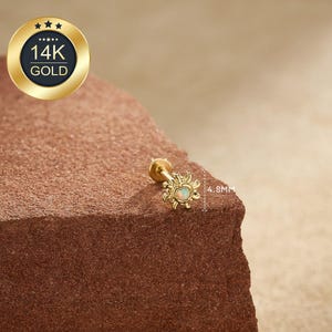 Może przedstawiać: Złota kolczyka w kształcie słońca z małym, owalnym, białym kamieniem opalu pośrodku. Kolczyk ma średnicę 4,8 mm. Obraz zawiera również złoty emblemat z napisem "14K GOLD".