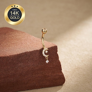 Puede incluir: Un piercing para el ombligo de color dorado con un diseño de barra curva. El anillo presenta un diseño de luna creciente y estrella con pequeñas piedras preciosas transparentes. Una gema colgante en forma de lágrima cuelga de la luna. El anillo se muestra sobre una superficie marrón texturizada.