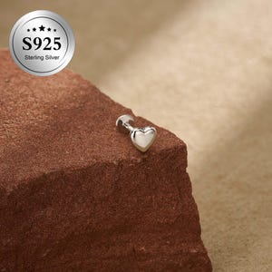 Può includere: Un orecchino a lobo a forma di cuore in argento sterling. L'orecchino è piccolo, con una finitura lucida, ed è posto su una superficie strutturata marrone-rossastra. L'immagine include anche un logo circolare con la scritta "S925 Sterling Silver."