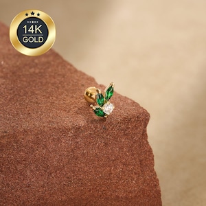 May include: A gold-colored earring with a leaf design, featuring three green marquise-cut stones and a central square-cut clear stone. The earring is set against a reddish-brown surface, with a gold seal that reads "14K GOLD".