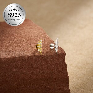 May include: Two feather-shaped earrings made of sterling silver, adorned with clear gemstones. One earring is gold-toned, the other silver-toned. They are displayed on a brown, textured surface. A silver emblem with the text "S925 Sterling Silver" is in the upper left corner.