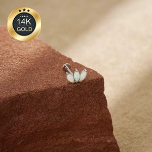 May include: A 14K gold body piercing stud with three opal gemstones. The stud is silver-colored and features a flat back. The opal stones are oval-shaped and have a milky white color. The stud is displayed on a brown, textured surface.