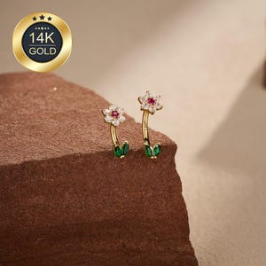 May include: A pair of gold earrings featuring a floral design with a red center stone and white accents. The earrings have a curved stem with green leaf details. The earrings are marked with "14K GOLD" in a gold circle.