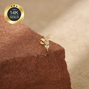 May include: A gold-coloured ear piercing stud featuring a leaf design, embellished with small, clear gemstones. The stud is set against a textured, reddish-brown surface. A gold emblem with "14K GOLD" is in the upper left corner.
