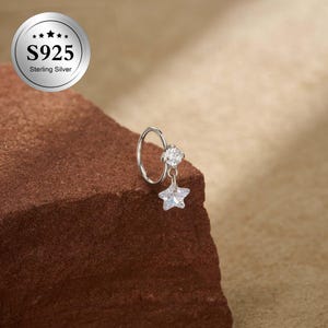 May include: A silver nose ring with a star-shaped charm. The ring has a clear, round gemstone and a star pendant. The image includes a silver emblem that reads "S925 Sterling Silver". The ring is displayed on a brown surface.