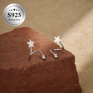 May include: A pair of sterling silver earrings with a star design. Each earring features a star at the top, a curved silver wire, and a small ball at the end. The earrings are displayed on a brown surface. The image also includes a silver circle with the text "S925 Sterling Silver."