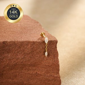 Könnte beinhalten: Ein goldfarbener 14K Gold Ohrpiercing-Stecker mit zwei herabhängenden, klaren, marquiseförmigen Zirkonia-Steinen. Der Stecker wird auf einer strukturierten, rotbraunen Oberfläche präsentiert. Ein goldfarbenes Emblem mit dem Text "14K GOLD" ist zu sehen.