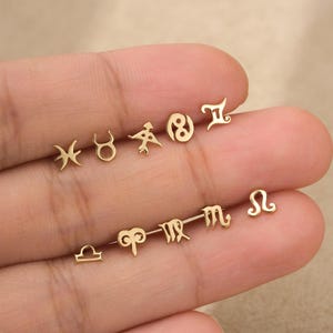 May include: A collection of small, gold-colored metal stud earrings, each shaped as a distinct zodiac sign symbol. The set includes individual pieces representing Pisces, Taurus, Sagittarius, Cancer, Gemini, Libra, Aries, Virgo, Scorpio, and Leo.